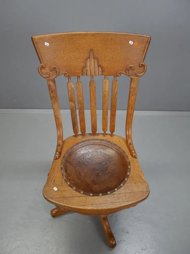 vintage revolving oak desk chair, slatted back w/ shaped crest, leather seat embossed wreath w/ helmet & sword. On 4-pt Image 1++